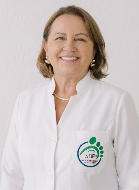 Diretoria / Regionais 16 Retrato em estúdio de Ana Adelia Vaine, presidente da SBP do Paraná. Ela é uma mulher de pele clara e cabelos curtos, que sorri para a câmera vestindo um jaleco branco com o logotipo oficial da Sociedade Brasileira de Podólogos bordado no bolso.