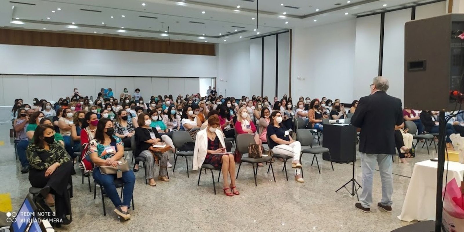 Grande auditório lotado com profissionais de podologia sentados em fileiras, a maioria mulheres usando máscaras de proteção, assistindo atentamente a uma palestra ministrada por um homem de costas no palco durante um evento oficial da SBP.