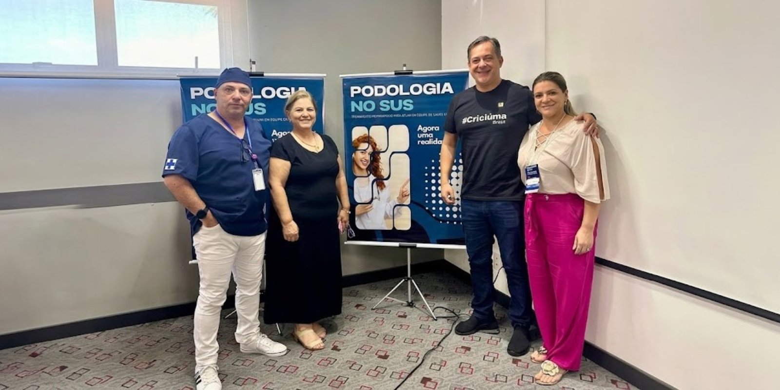 Quatro pessoas posando em pé e sorrindo para a foto, incluindo um profissional de saúde de pijama cirúrgico azul e um homem usando uma camiseta preta com a hashtag Criciúma. Atrás do grupo, destacam-se dois banners azuis com o título "Podologia no SUS: Agora uma realidade