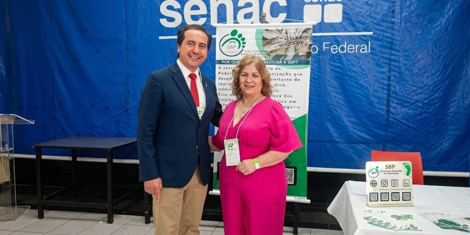 Um homem sorridente de terno azul e gravata vermelha ao lado de uma mulher de macacão rosa que usa um crachá da SBP. Eles posam em frente a um banner informativo com o título "Por que se associar à SBP?" e um painel azul de fundo com o logotipo do Senac. À direita, há uma mesa com panfletos e um display com QR codes da Sociedade Brasileira de Podólogos.